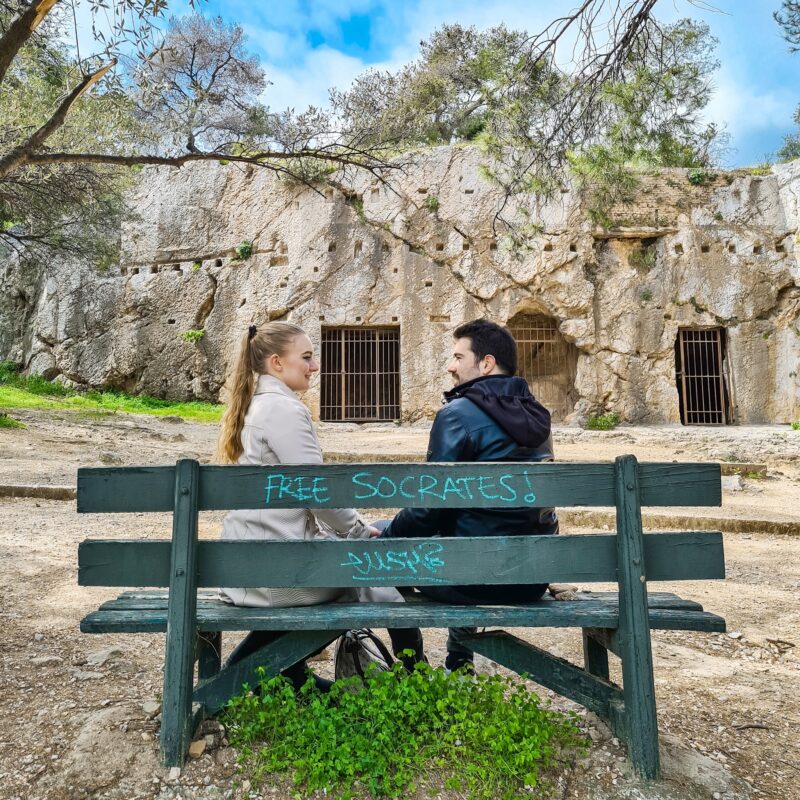 Travel Couple posing on bench in front of The Prison of Socrates in Athens, Greece