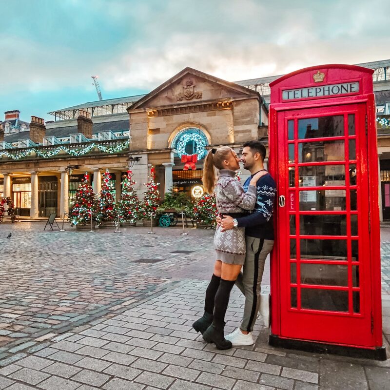 Travel Couple posing with Christmas Decorations and iconic red phone booth at Covent Garden in London